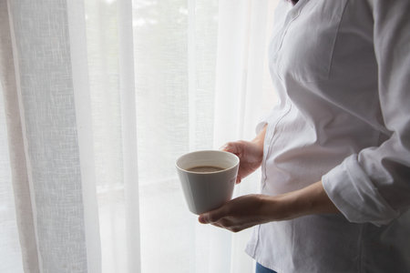 young woman holding a cup of coffee on curtain windows texture background, side view.