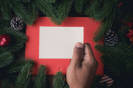 Human hand holding blank note paper on Christmas decorations background with pine leaf frame, view from above table.の写真素材