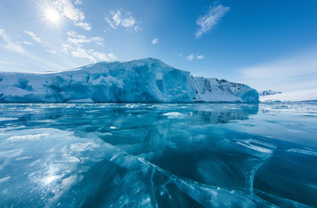 Transparent ice with detailed cracks on the Greenlandic surface, alongside a large, smooth iceberg under a clear sky with intense sunlightの素材