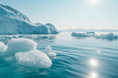 Icebergs and smaller ice formations float on the clear waters of Greenland under a blue sky, with sunlight reflecting on the waters surfaceの素材