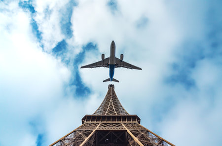 A passenger airplane captured from below as it flies directly over the Eiffel Tower against a cloudy blue skyの素材