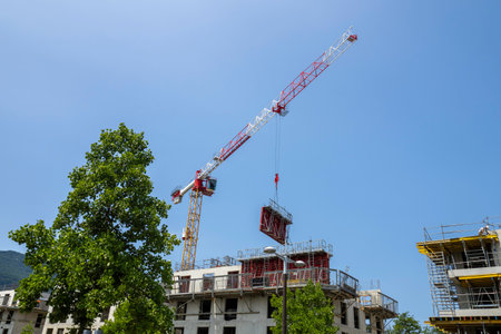 Tall construction crane lifting materials at a building site against a clear blue skyの写真素材