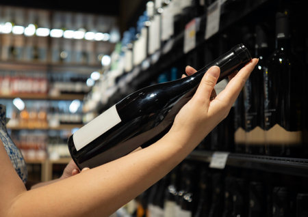 Close-up of a hand holding a wine bottle in a store aisle with shelves of wine bottles in the backgroundの写真素材