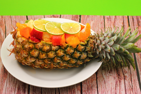 Thai style fruit salad inside a pineapple with a green background on a wood table topの写真素材