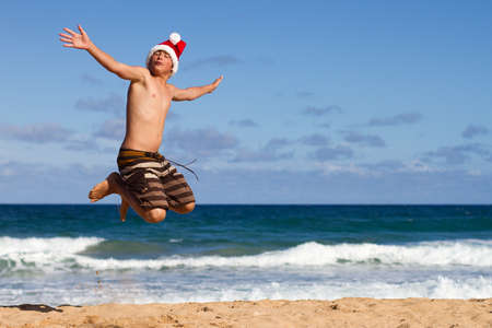 A Teen Boy Wearing a Santa Hat Jumping on the Beach in Hawaiiの写真素材
