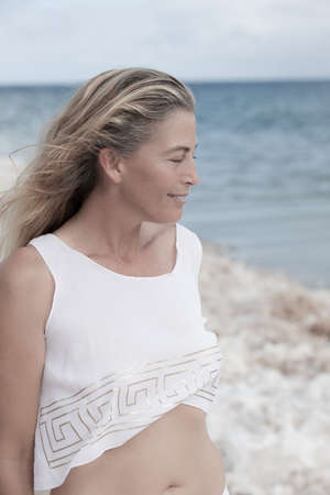 Beautiful woman with long hair in her Forties wearing a white sheer top  on the beachの写真素材