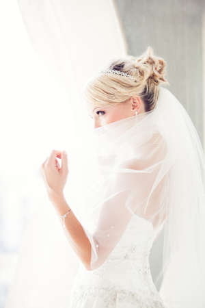 Gorgeous smiling blonde bride  in front of a Penthouse window at a modern hotel peaking over her veilの写真素材