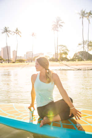 Sunny morning sun flare moment of a pretty young woman in SUP Yoga practice in twist poseの写真素材