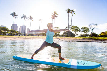 Sunny morning sun flare moment of a pretty young woman in SUP Yoga practice in warrior poseの写真素材