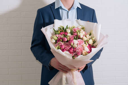 young blond man in a blue suit holds in hand a giant bouquet of white-pink roses and eustoma on the light wall backgroundの写真素材