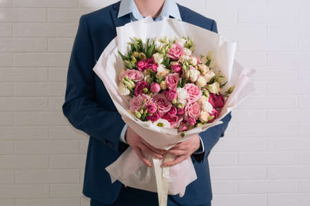 young blond man in a blue suit holds in hand a giant bouquet of white-pink roses and eustoma on the light wall backgroundの写真素材