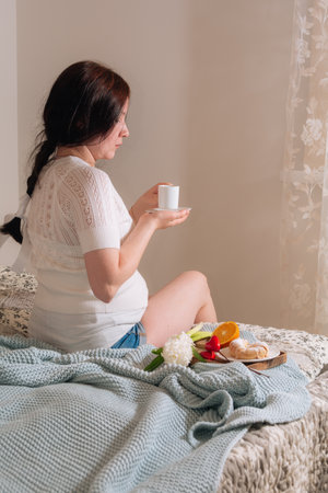 Pretty brunette pregnant girl in white clothes holding cup of coffee and enjoying in bed room during morning timeの写真素材