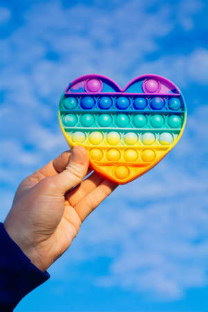 colorful antistress sensory toy fidget push pop it in man hand on blue sky background. rainbow pop it heart on skyの写真素材