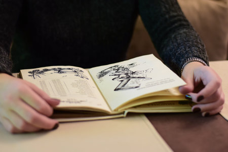 unrecognizable young woman with dark nail polish holding open menu book in gentle hands.