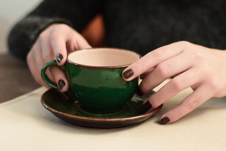 young female hands with dark nail polish holding beautiful dark green coffee cup on platter with hot green teaの写真素材