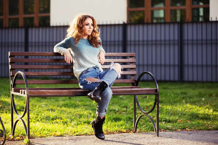Happy young fashion woman with long curly hairs sitting on bench in city parkの写真素材