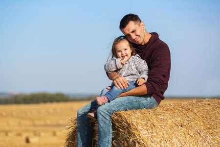 Happy father and 2 year old girl sitting on hay bale in harvested fieldの写真素材