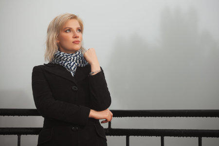 Young fashion woman in black coat leaning on handrail in a fog outdoorの写真素材