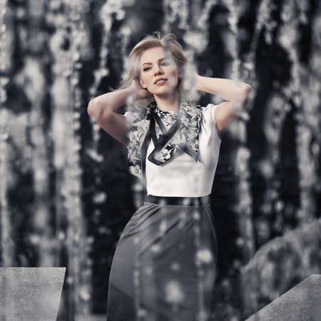 Happy young fashion blond woman walking in city street against a fountain. Stylish female model in white blouse and grey skirt outdoorの写真素材
