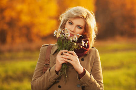 Happy young blond woman with flowers walking outdoor Stylish fashion model wearing classic beige coat and burgundy scarfの写真素材