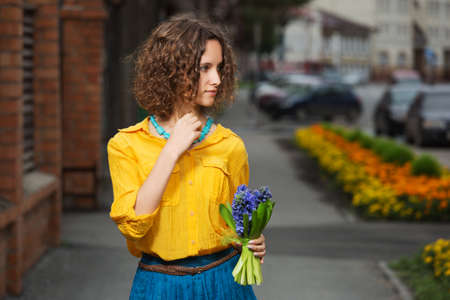 Young fashion woman with bouquet of flowers walking in city street Stylish female model wearing yellow shirt and blue skirtの写真素材
