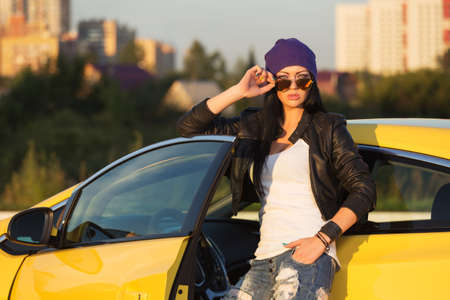 Young fashion hipster woman in sunglasses leaning on her car  Stylish female model in black leather jacket purple beanie and ripped jeansの写真素材