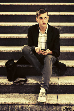 Young business man with cell phone sitting on steps Stylish male model in black blazer and gray...