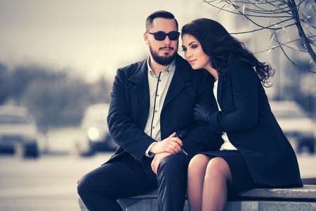 Happy young fashion couple in love sitting on bench  Stylish trendy man and woman in classic black coatsの写真素材