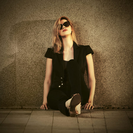 Sad young fashion woman in sunglasses sitting at the wall on city street Stylish female model wearing dark blue jacket and jeans の写真素材