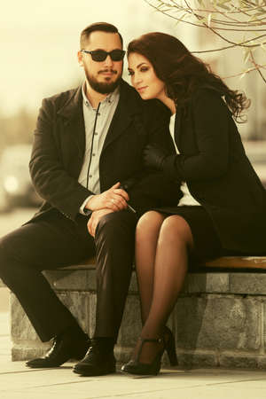 Happy young fashion couple in love sitting on bench  Stylish trendy man and woman in classic black coatsの写真素材