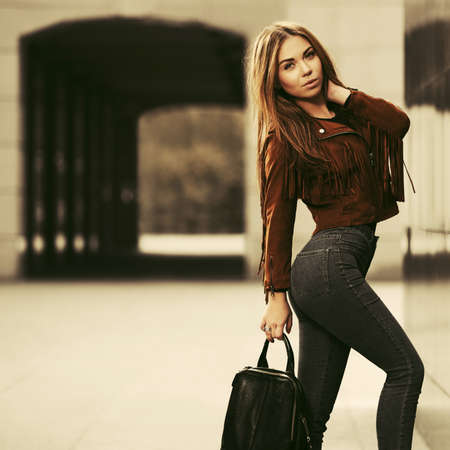 Young fashion woman with handbag walking on city street Stylish female model in brown leather fringe suede jacket and dark blue jeansの写真素材