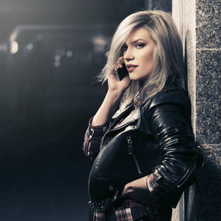 Young fashion blonde woman talking on cell phone on city street at night Stylish female model in black leather jacketの写真素材