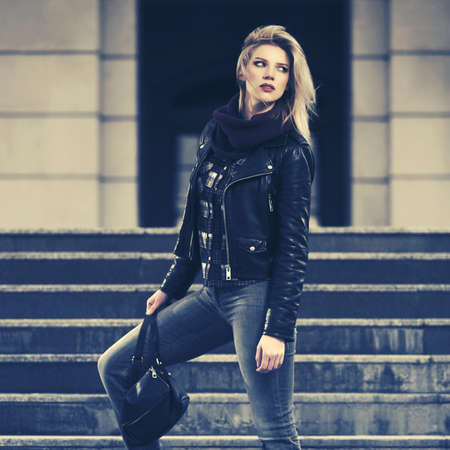 Young fashion blonde woman with handbag walking on city street  Stylish female model in black leather jacket and snood scarfの写真素材