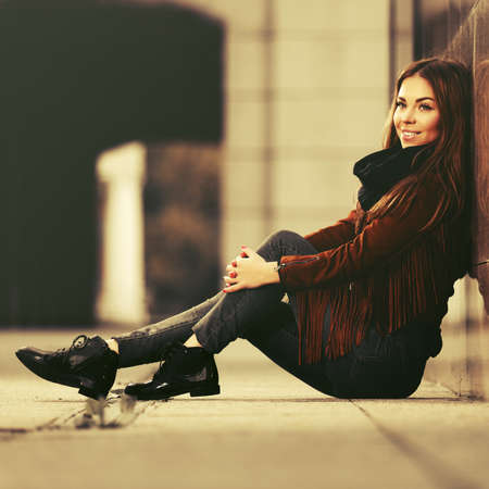 Happy young fashion woman sitting on city street sidewalk  Stylish female model in brown leather fringe suede jacket and black scarfの写真素材