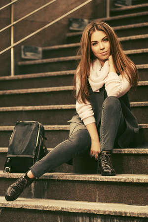 Happy young fashion woman with handbag sitting on steps Stylish female model in gray sleeveless coat and light pink sweaterの写真素材