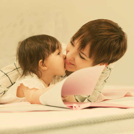 Happy young mother and little baby girl reading a book on bedの写真素材
