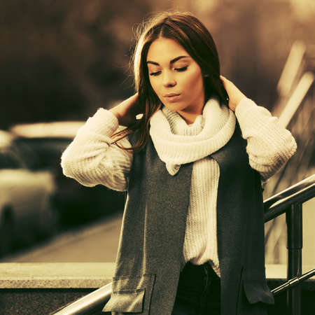 Sad young fashion woman leaning on railing Stylish female model in gray sleeveless coat and light pink sweaterの写真素材