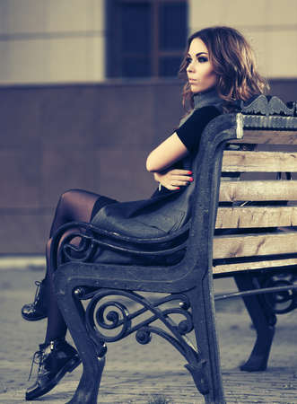 Sad young fashion woman with long curly hairs sitting on bench  Stylish female model in long sleeveless coatの写真素材