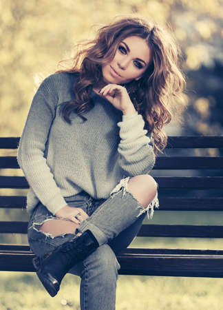 Happy young woman sitting on bench in city park Stylish fashion model in ripped jeans and pulloverの写真素材