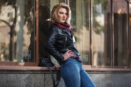 Young fashion blonde woman leaning on window  Stylish female model in black leather jacket and snood...