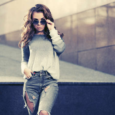 Happy young woman with long curly hairs in sunglasses walking on city street Stylish fashion model in gray pullover and ripped jeansの写真素材
