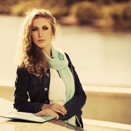Young blonde business woman with long curly hairs leaning on parapet Stylish fashion model in denim jacket and light green scarfの写真素材