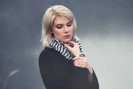 Sad young fashion woman walking in a fog outdoor Stylish female model in black coat and striped scarfの写真素材
