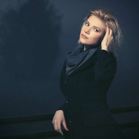 Happy young fashion woman leaning on railing in a fog  Stylish female model in classic black coat and gray scarfの写真素材