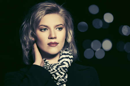 Young fashion blonde woman walking on city street at night Stylish female model in classic black coat and striped scarfの写真素材