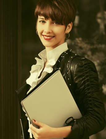 Happy young fashion business woman with file folder walking on city street Stylish trendy model with pixie hair in black leather jacket and white blouseの写真素材