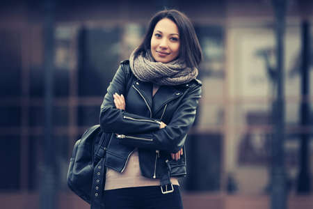 Young fashion woman with backpack walking on city street Stylish female model in black leather jacket and gray snood scarfの写真素材