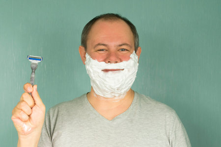 a man in shaving foam on his face is about to shave and holds a razor in his hand on a green backgroundの写真素材