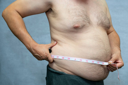 a fat man measures his waist with a bare stomach and chest, on a gray background