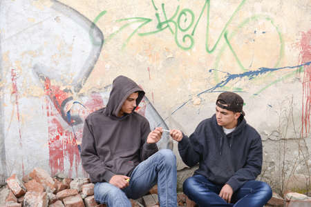 two street hooligans or rappers standing against a graffiti painted wall and sharing a cigaretteの写真素材
